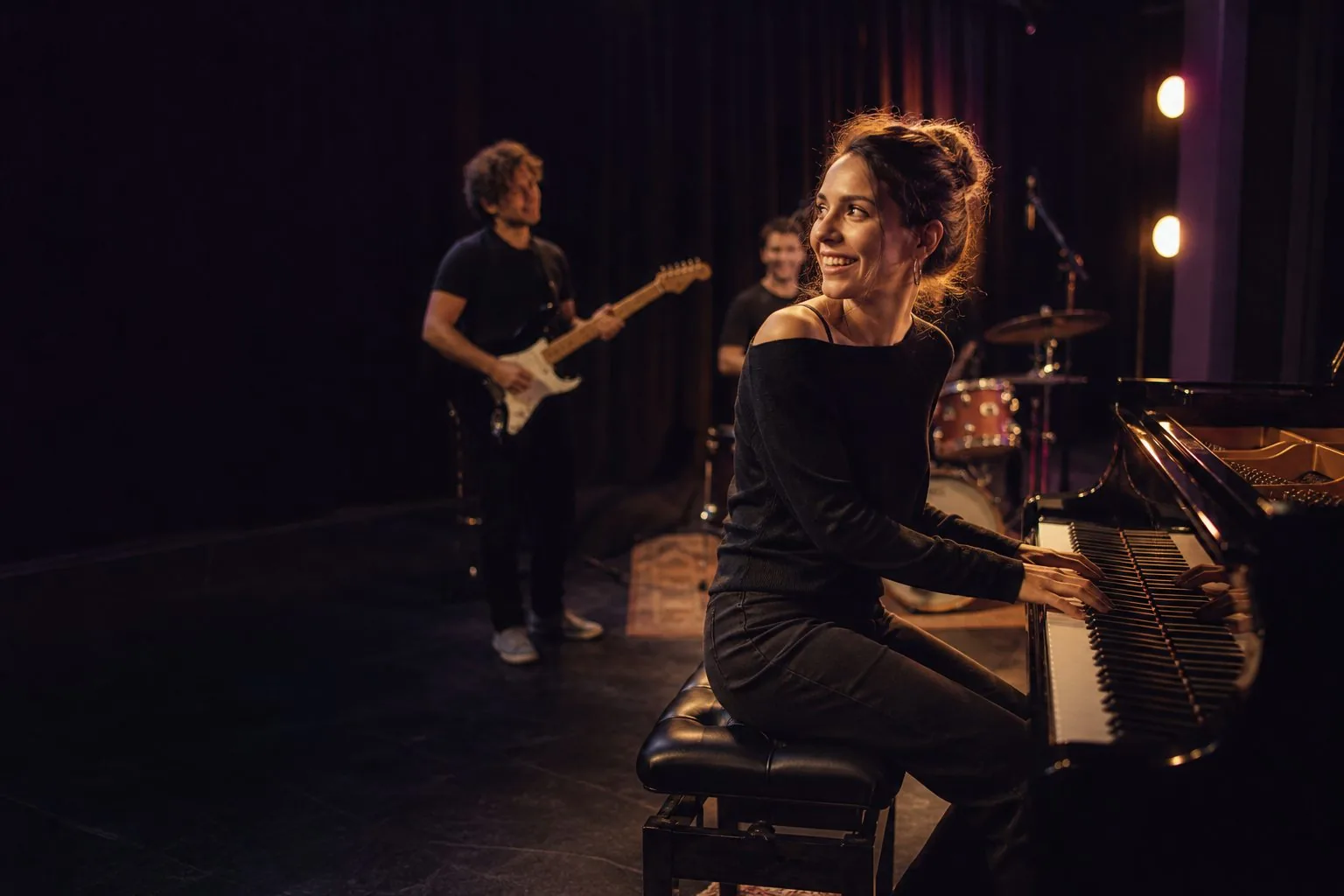 Une pianiste répète avec un guitariste et une batteuse dans une petite salle de scène